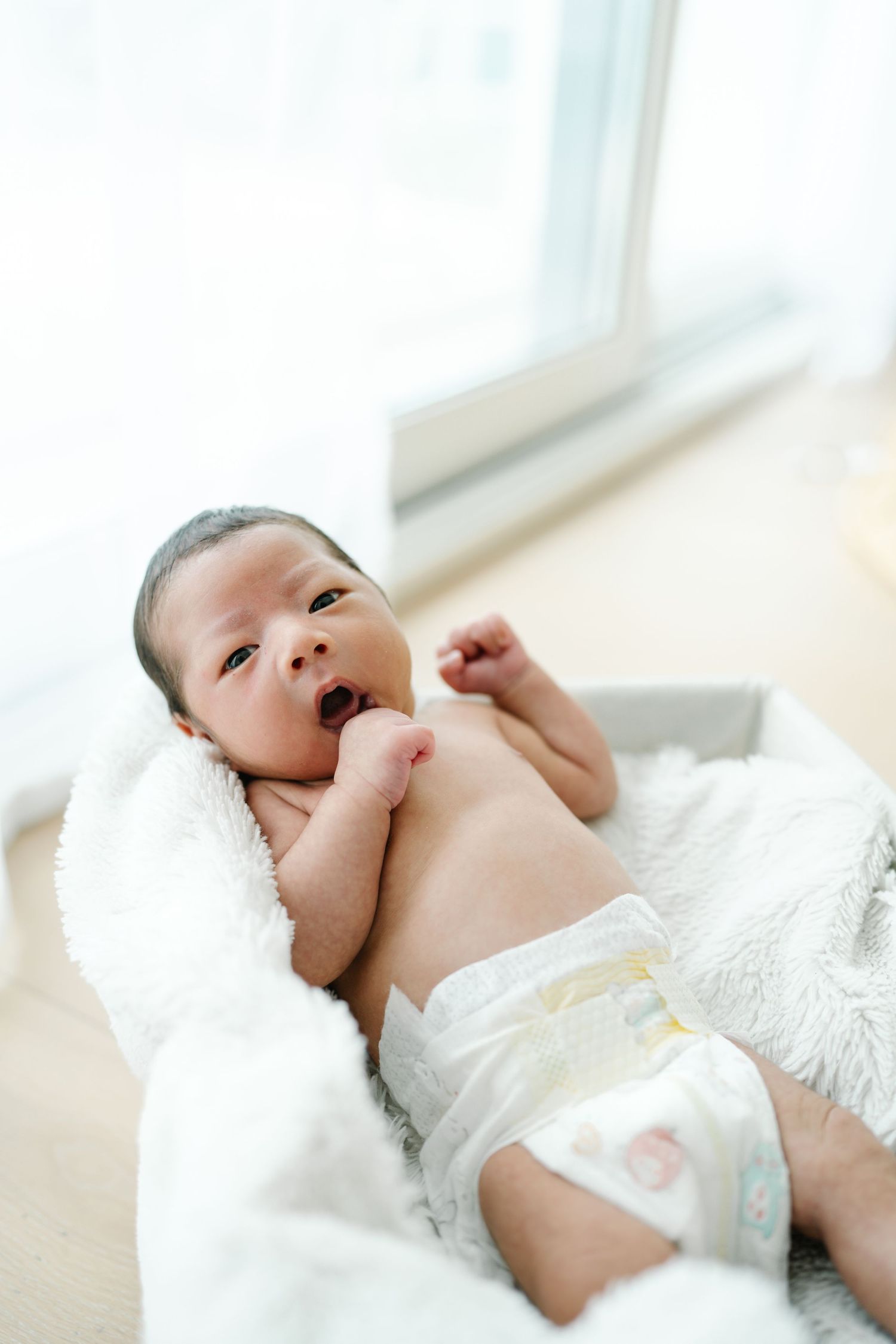 Alert newborn baby lying on soft white blanket near window.