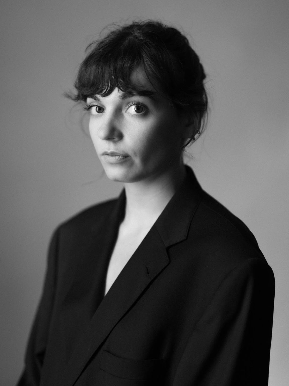 Black and white portrait of a young woman with bangs, hair up, wearing a black blazer against a neutral background.