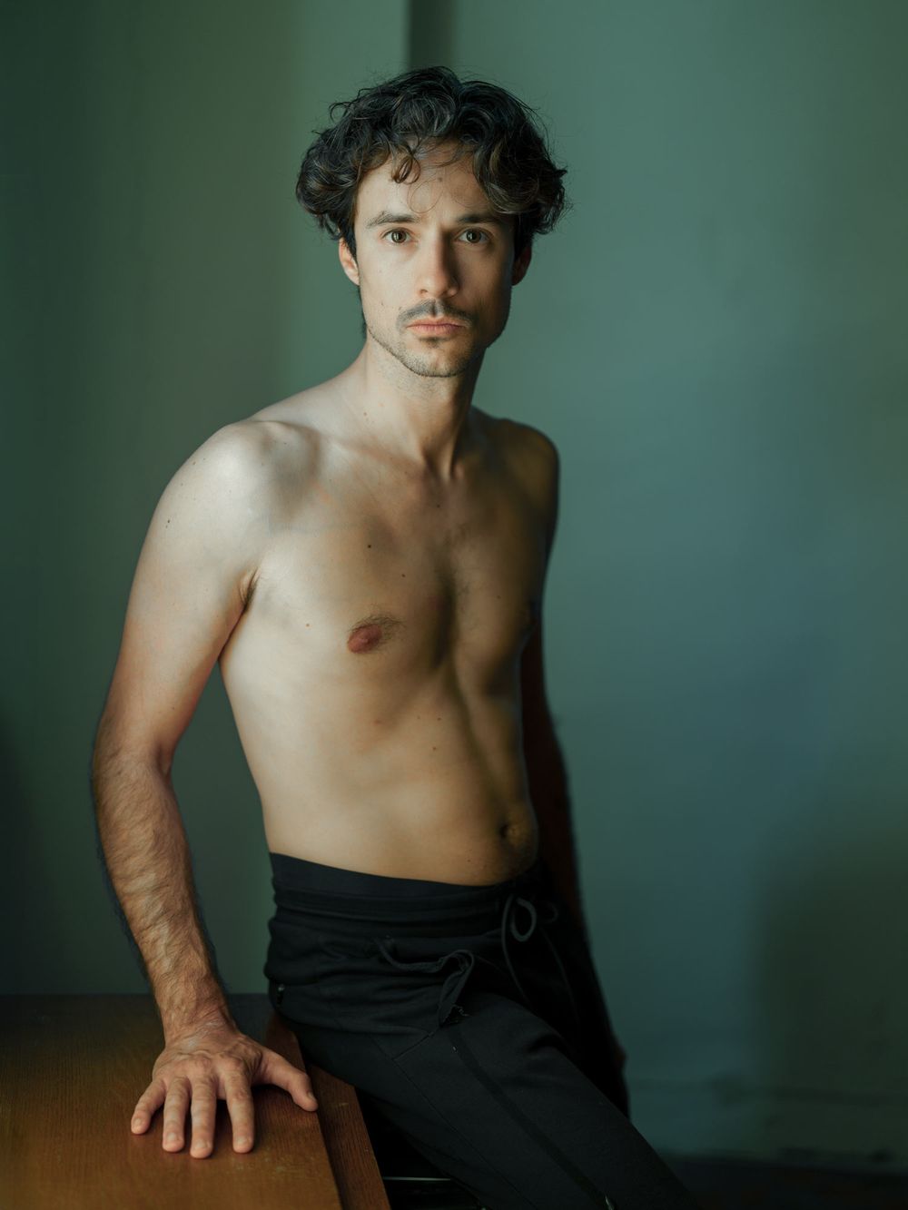 Shirtless young man with dark curly hair posing confidently in black pants against a muted green wall.