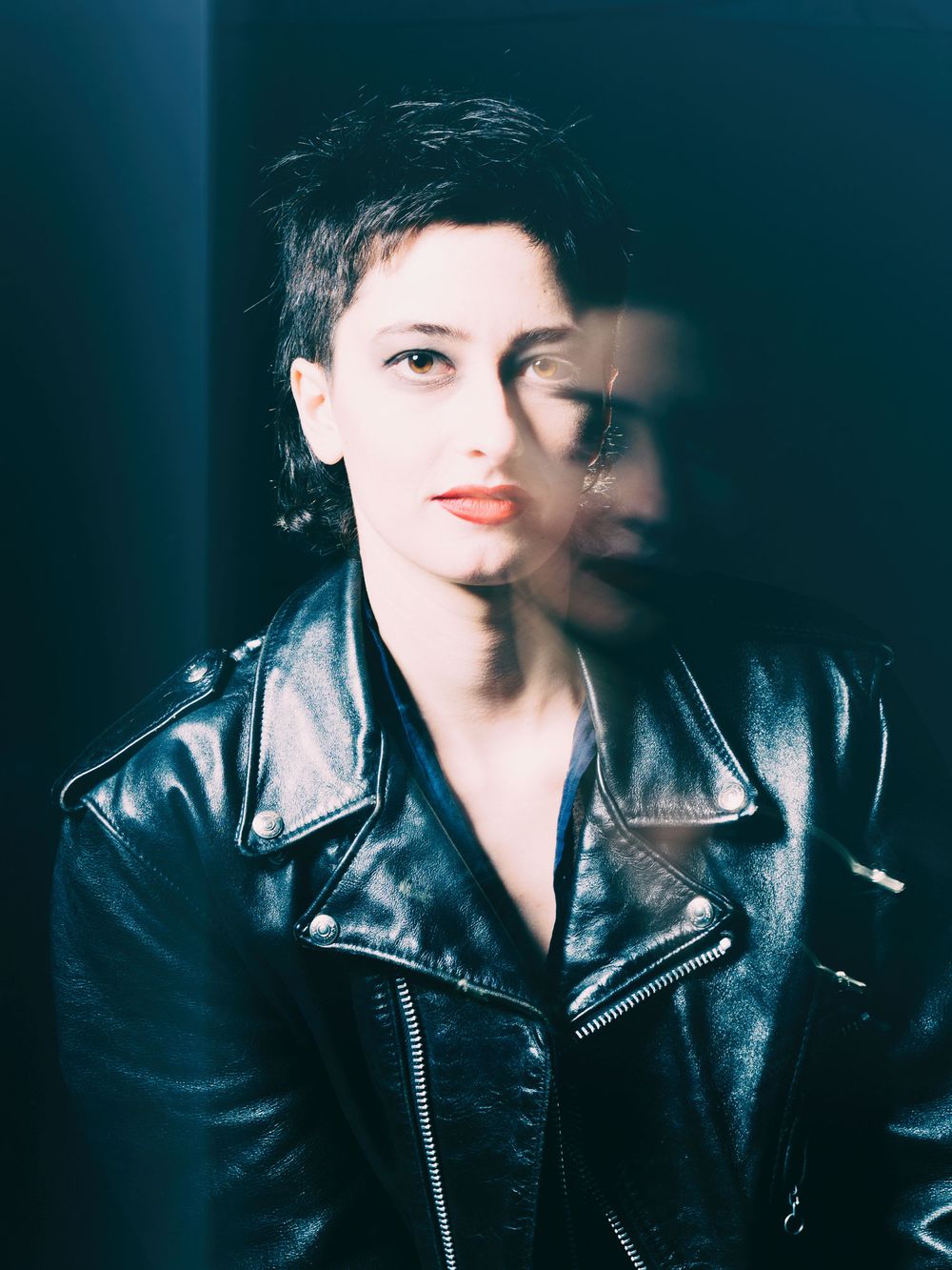 Woman in black leather jacket with short dark hair poses against dark background with dramatic reflection effect.