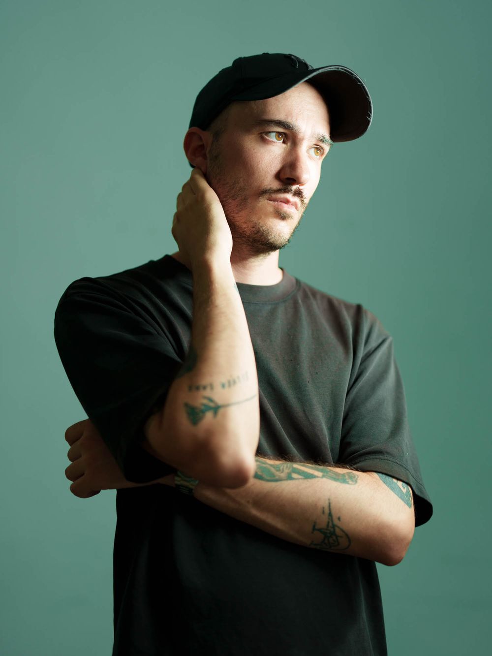 Young man with tattoos and black cap poses thoughtfully against teal background in dark t-shirt.