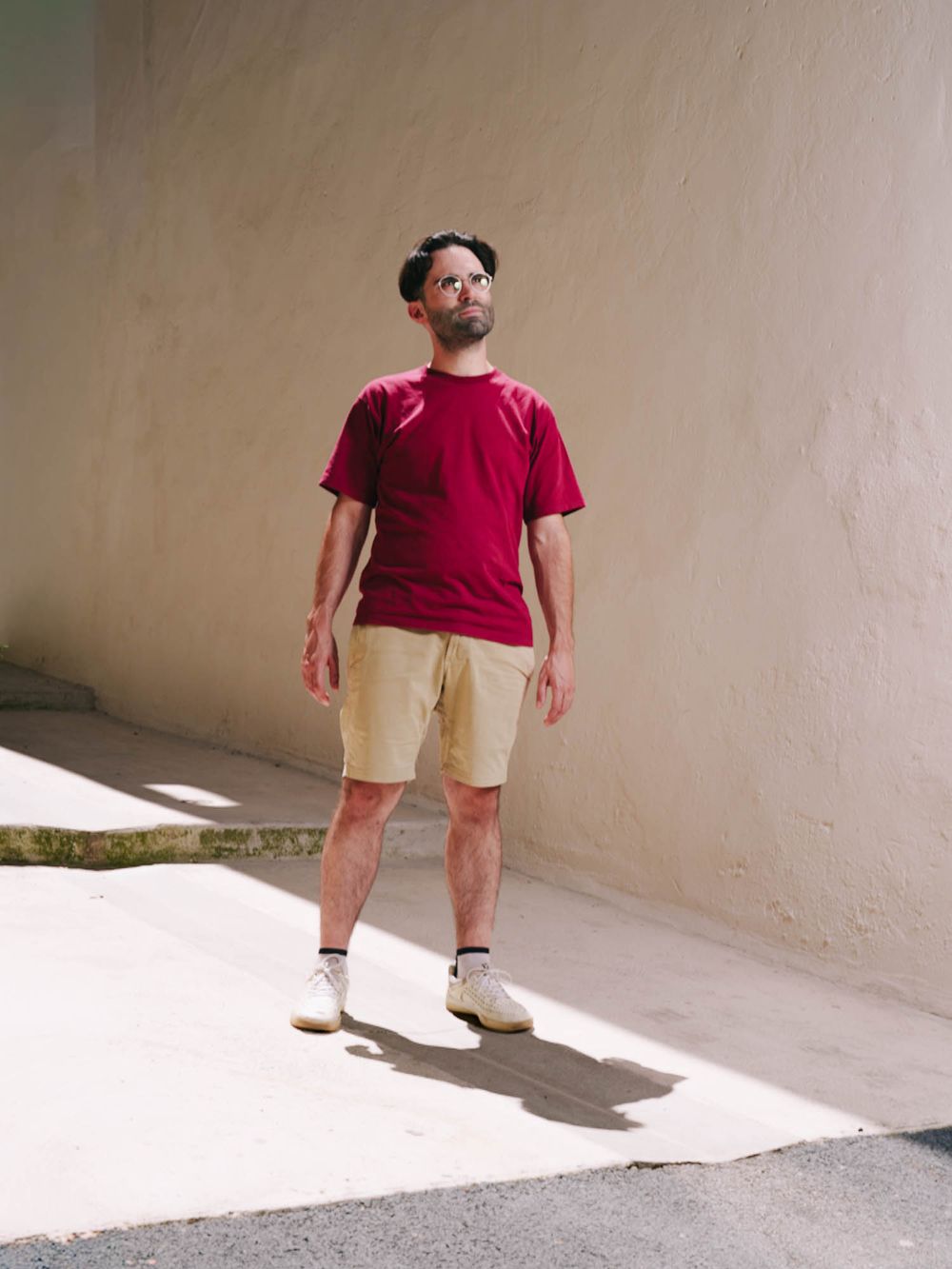 Man in red t-shirt and khaki shorts standing outdoors against a beige wall in sunlight.