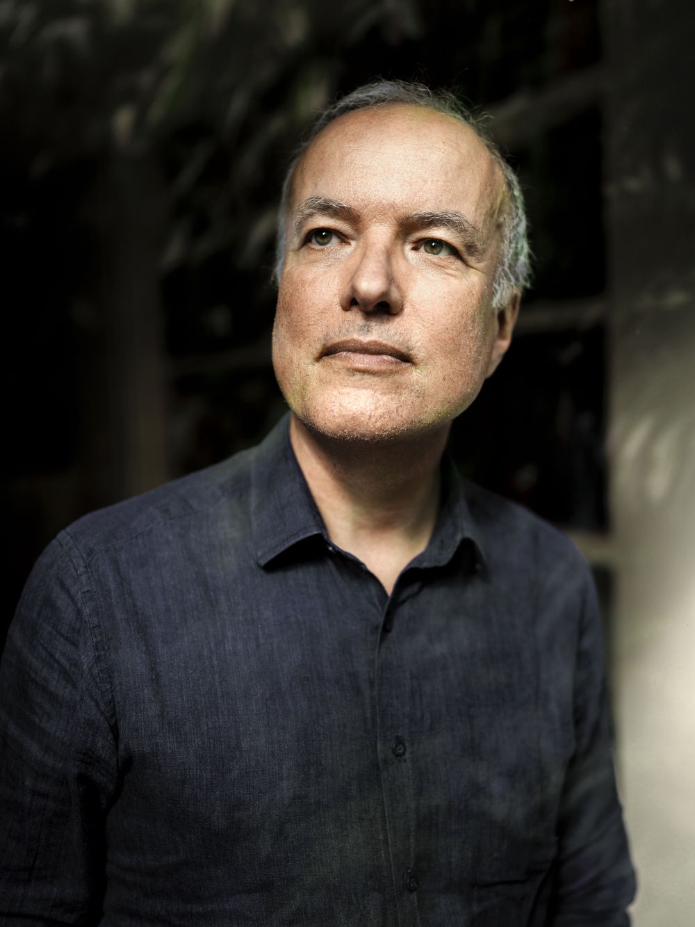Portrait of a middle-aged man with gray hair and a calm expression, wearing a dark linen button-up shirt against a dim, leafy background.