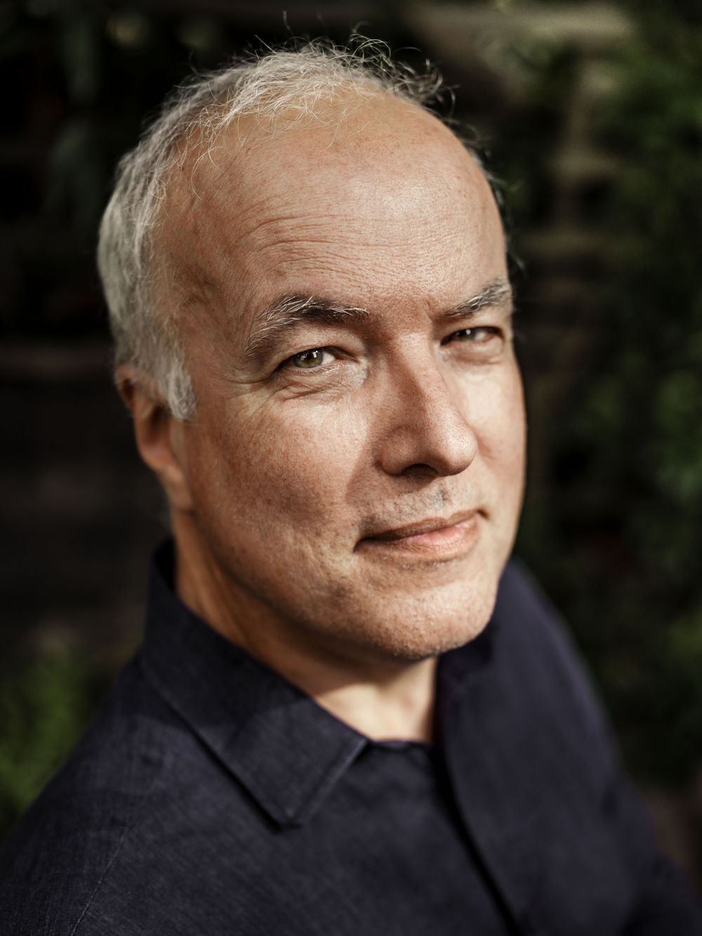 Portrait of a smiling middle-aged man with gray hair and a dark button-up shirt, photographed outdoors against a blurred green background.