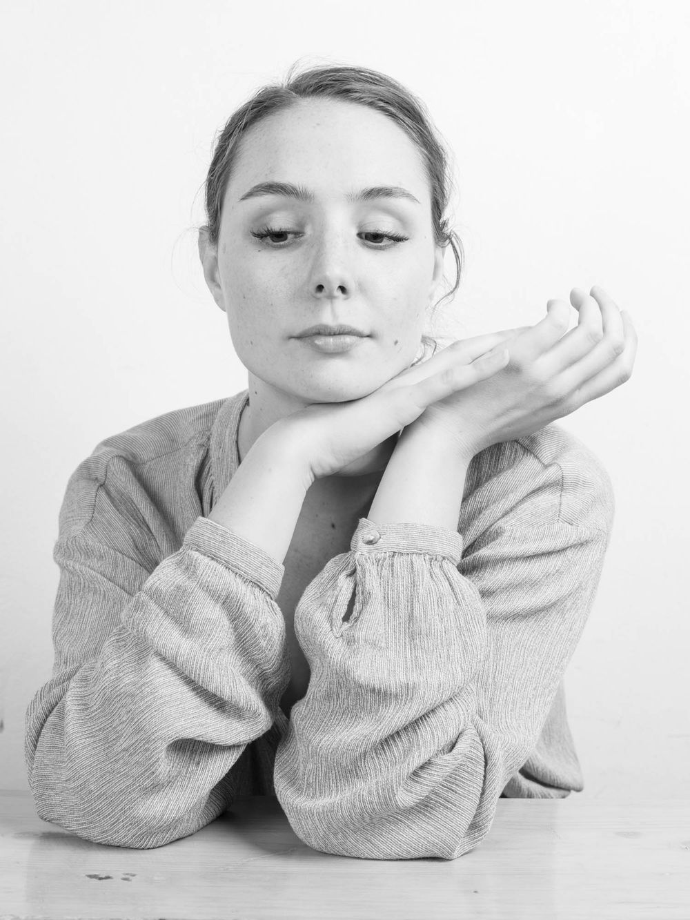 Black and white portrait of a young woman resting her chin on folded hands, wearing a cozy sweater.