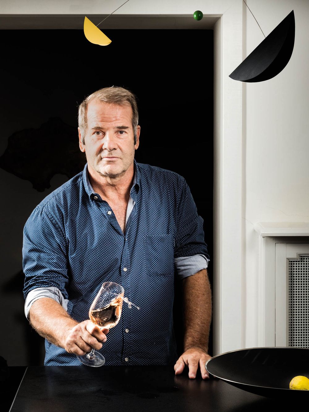 Man in blue shirt holding a wine glass at a dark table with colorful mobile art hanging above.