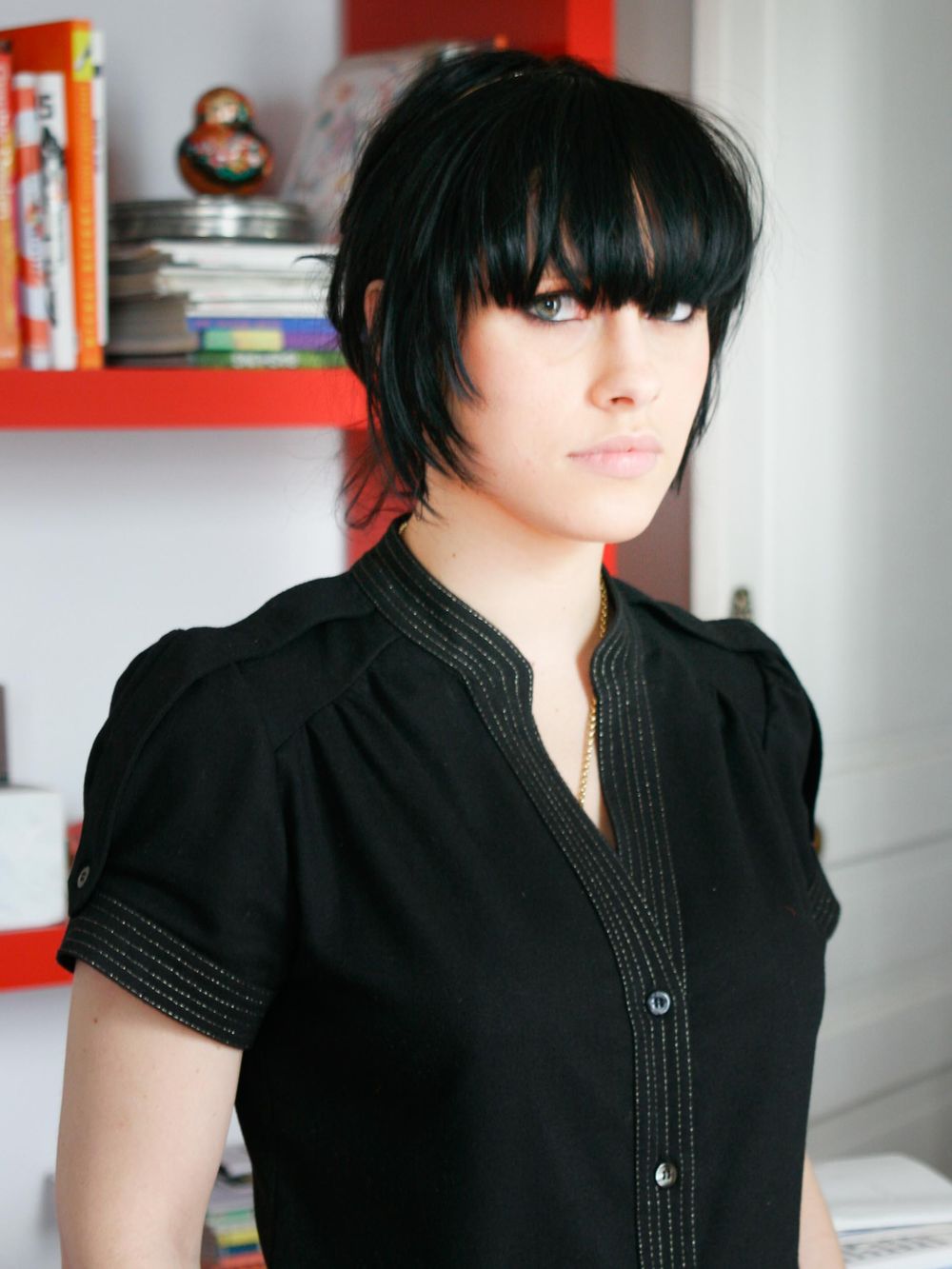 Person wearing a black button-up short sleeve top with dark bangs hairstyle standing against white wall with red shelving.
