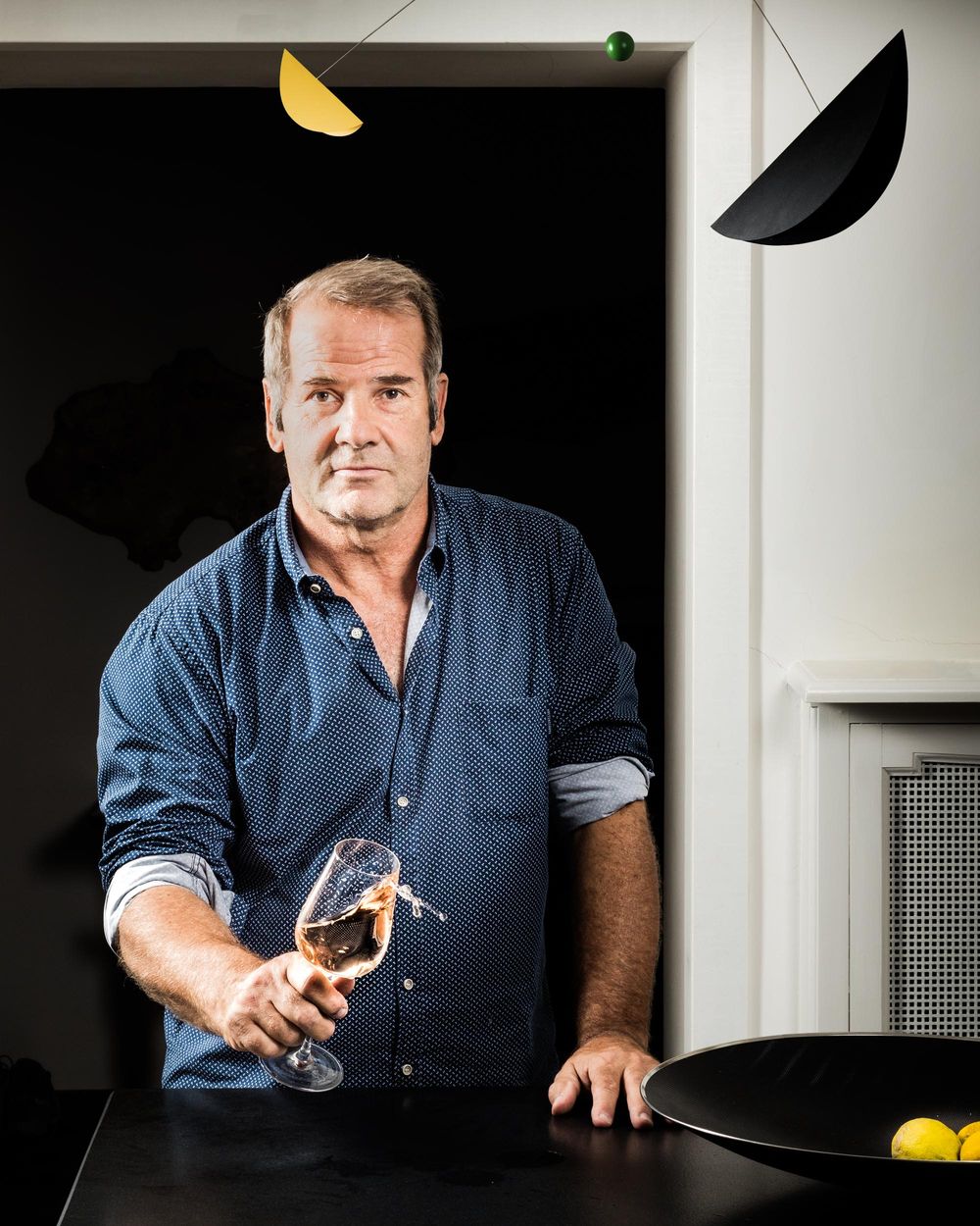 Chef in a navy button-down shirt stands at dark counter with artistic lighting and geometric wall decor.