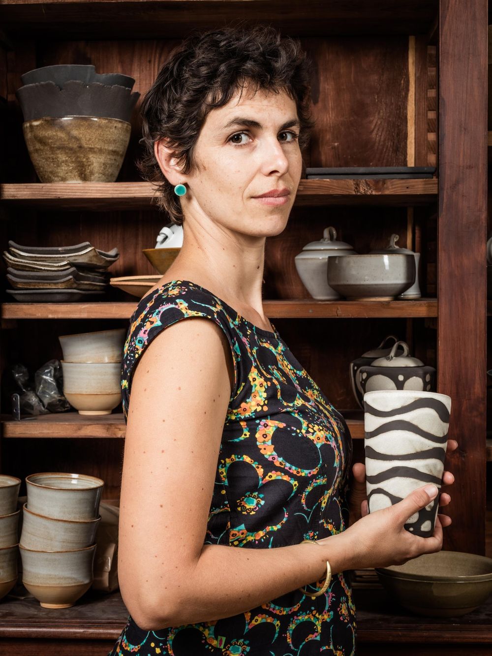 Stand-out portrait against a rustic backdrop of handmade pottery on wooden shelves, wearing a colorful patterned dress.