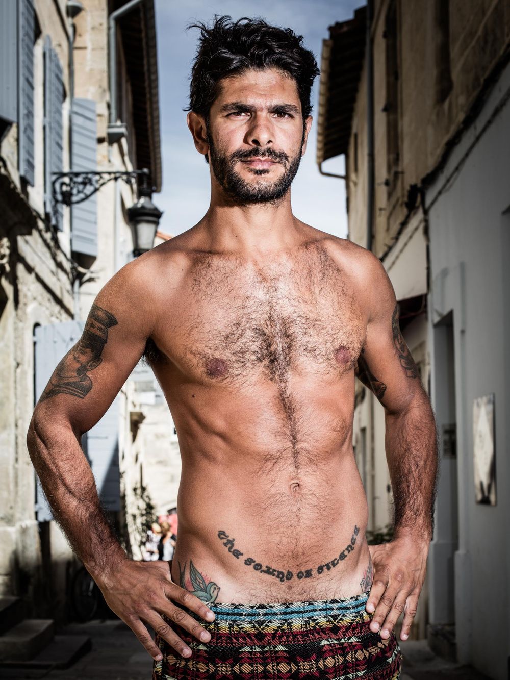 Shirtless athletic man with tattoos stands confidently in a narrow European alleyway during daytime.