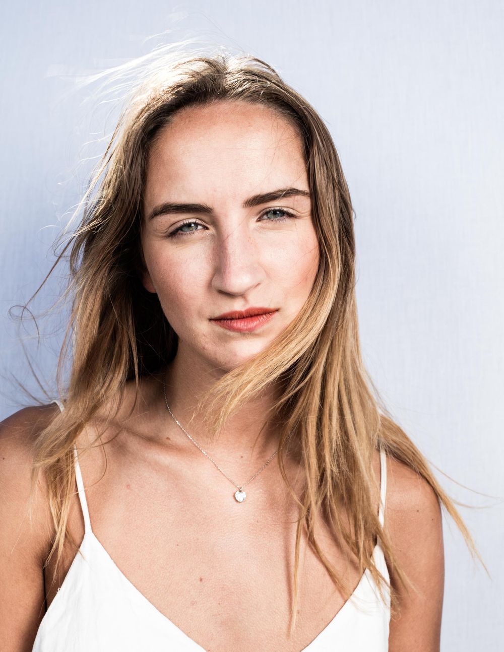 Portrait of someone with long blonde hair wearing a white top and delicate necklace against a light background.