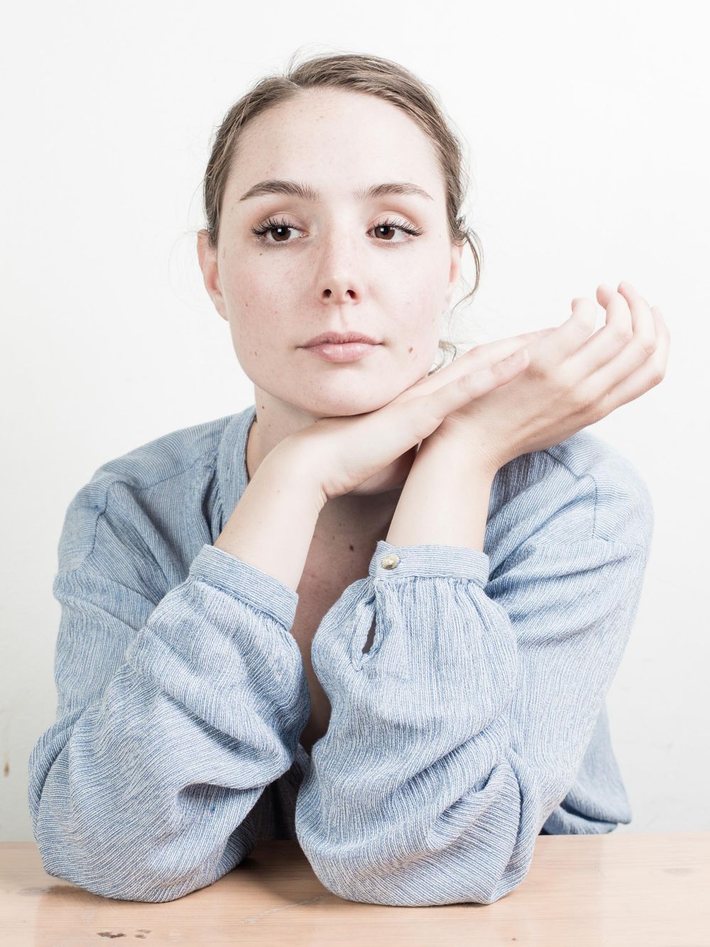 Person in a soft blue sweater rests their chin on folded hands while looking at camera against white background.