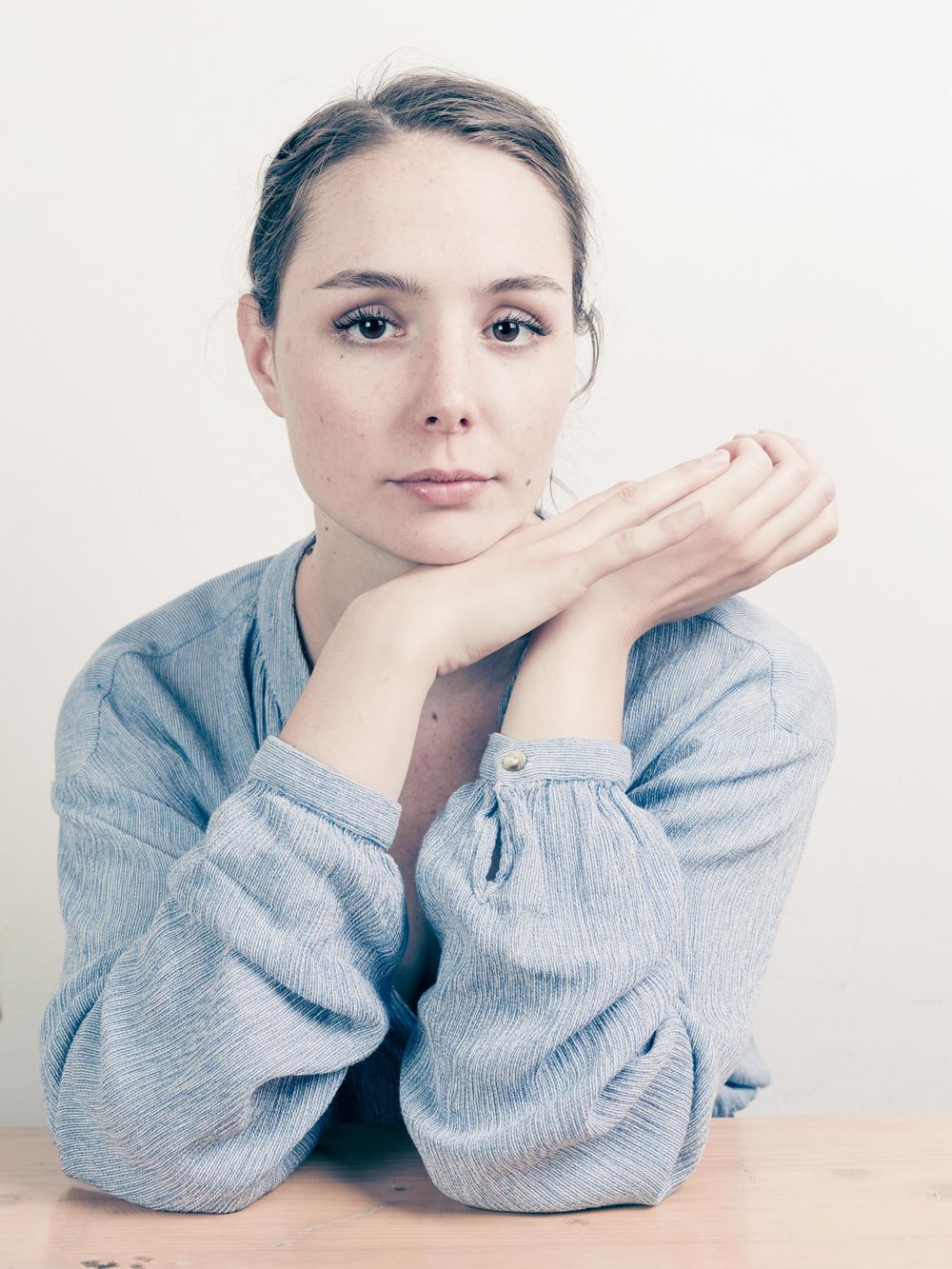 Artistic portrait with a light gray background showing someone wearing a chambray shirt with hands resting under their chin.