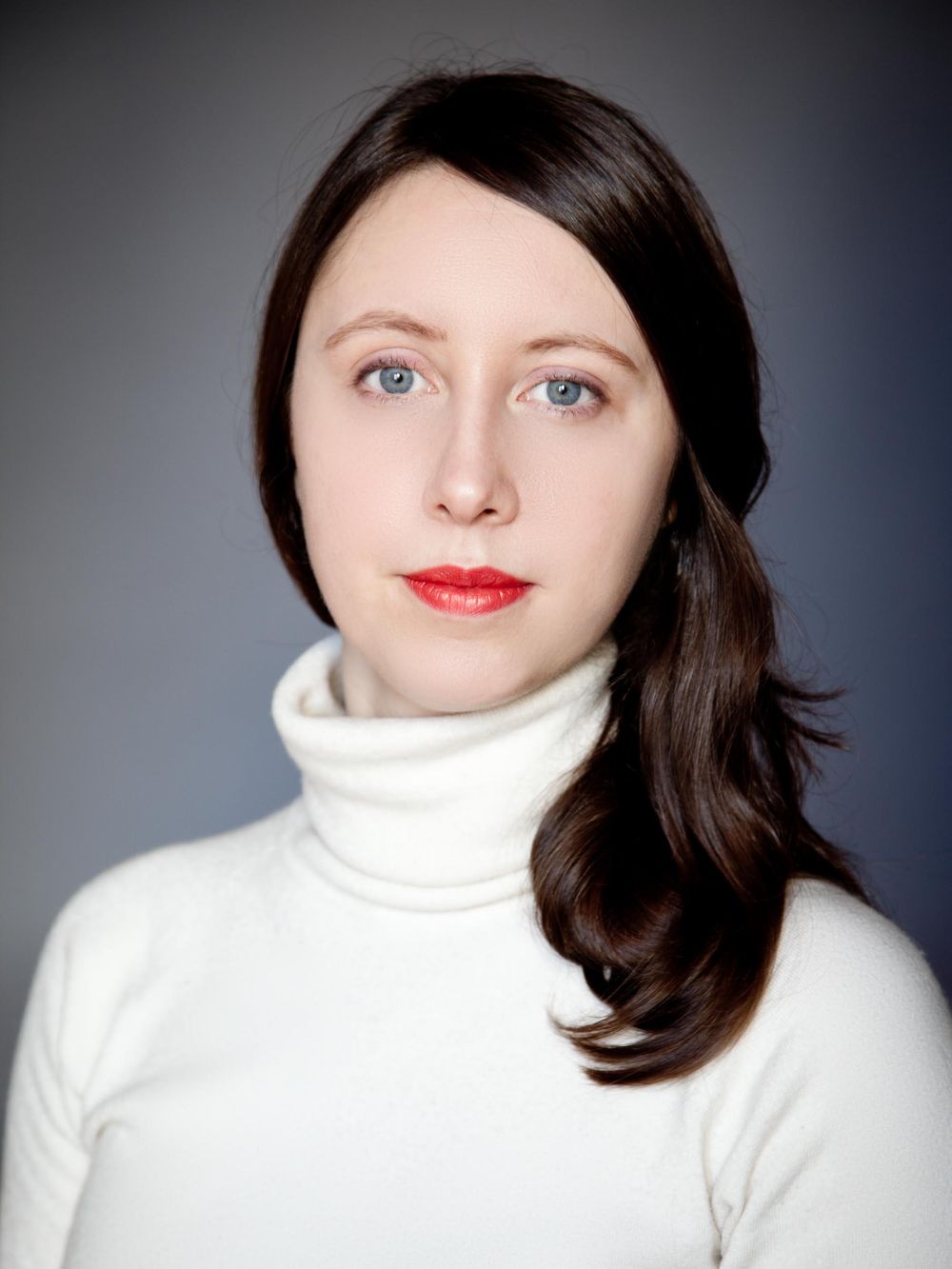 Portrait photo against gray background showing person with dark hair wearing white turtleneck sweater and bright red lipstick.