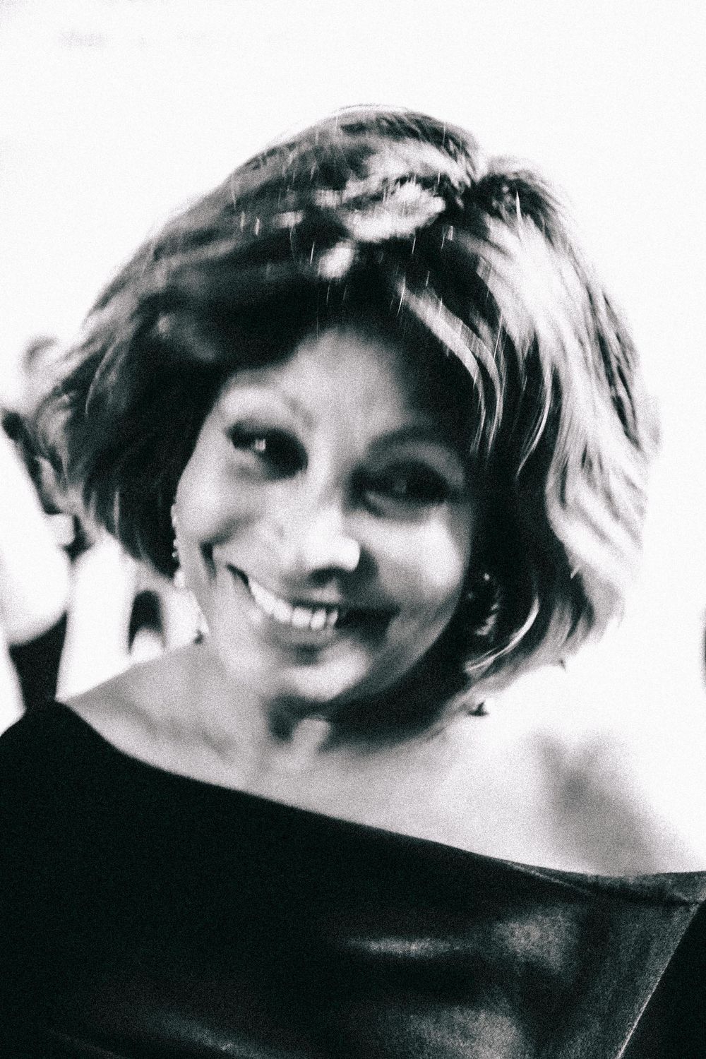 Black and white portrait showing someone with a warm smile and short wavy hair wearing an off-shoulder top.