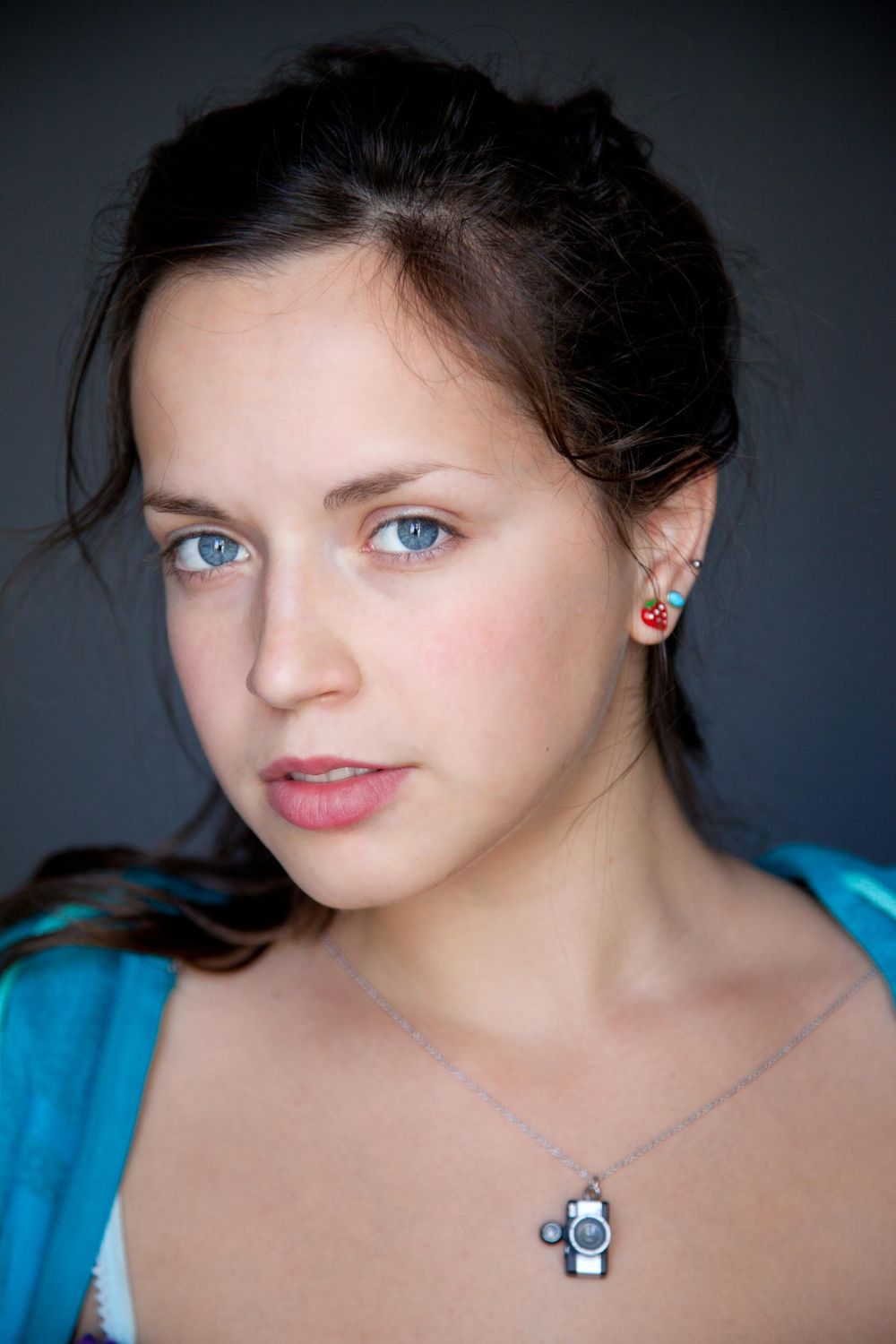 Portrait with bright blue eyes wearing a turquoise top and delicate necklace against dark background.