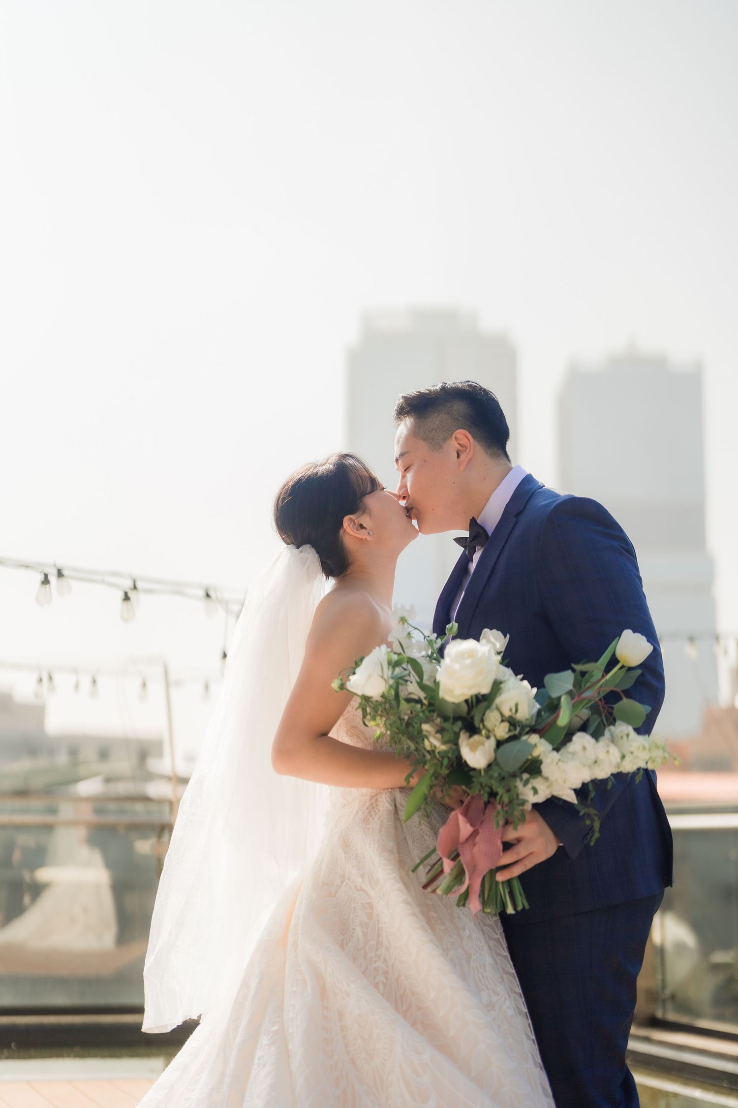 A couple in wedding attire share a kiss on a rooftop holding a bouquet of white flowers against a city skyline backdrop.