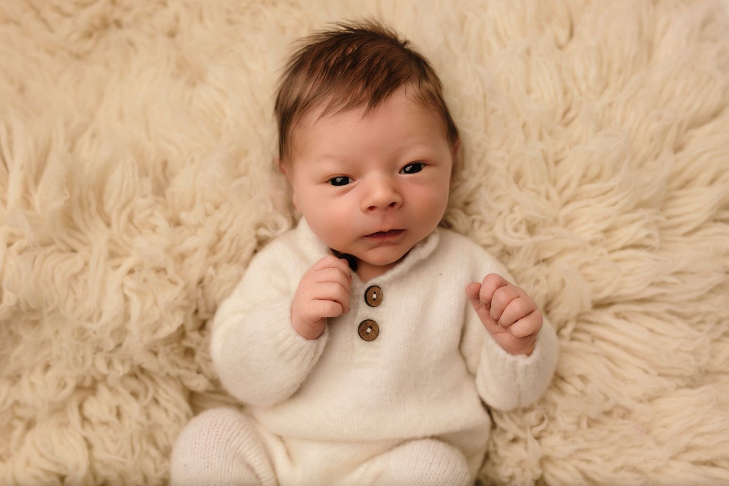 Charlottesville newborn photographer, baby boy in cozy white knit suit and lying against a wool rug looking quizzically at the camera.
