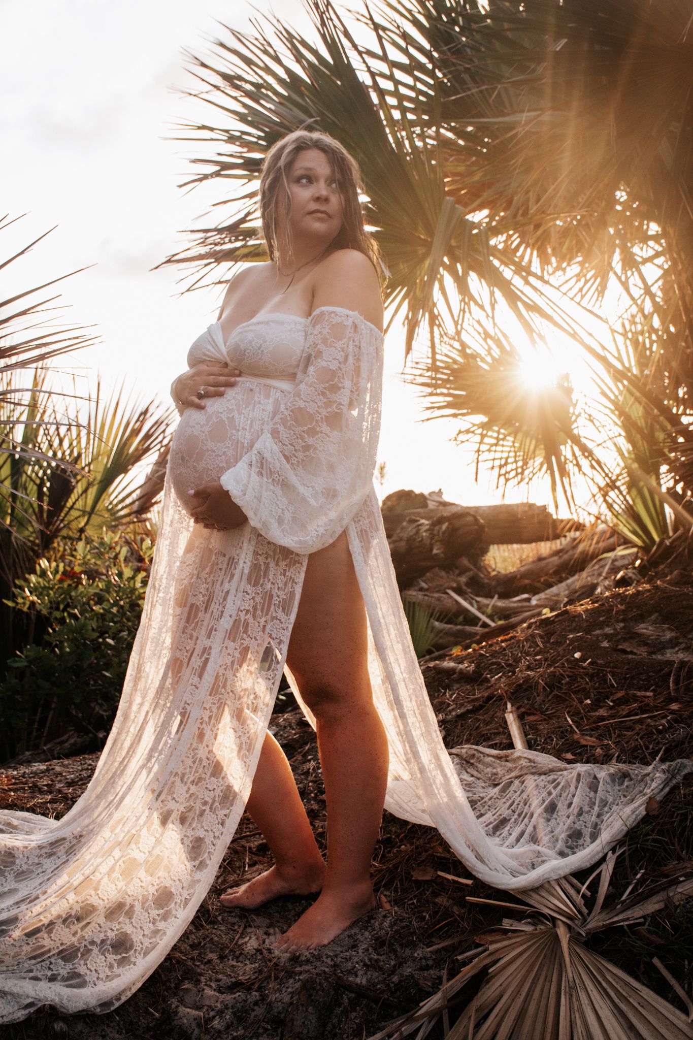 Maternity portrait with white lace robe at golden hour among palm trees.
