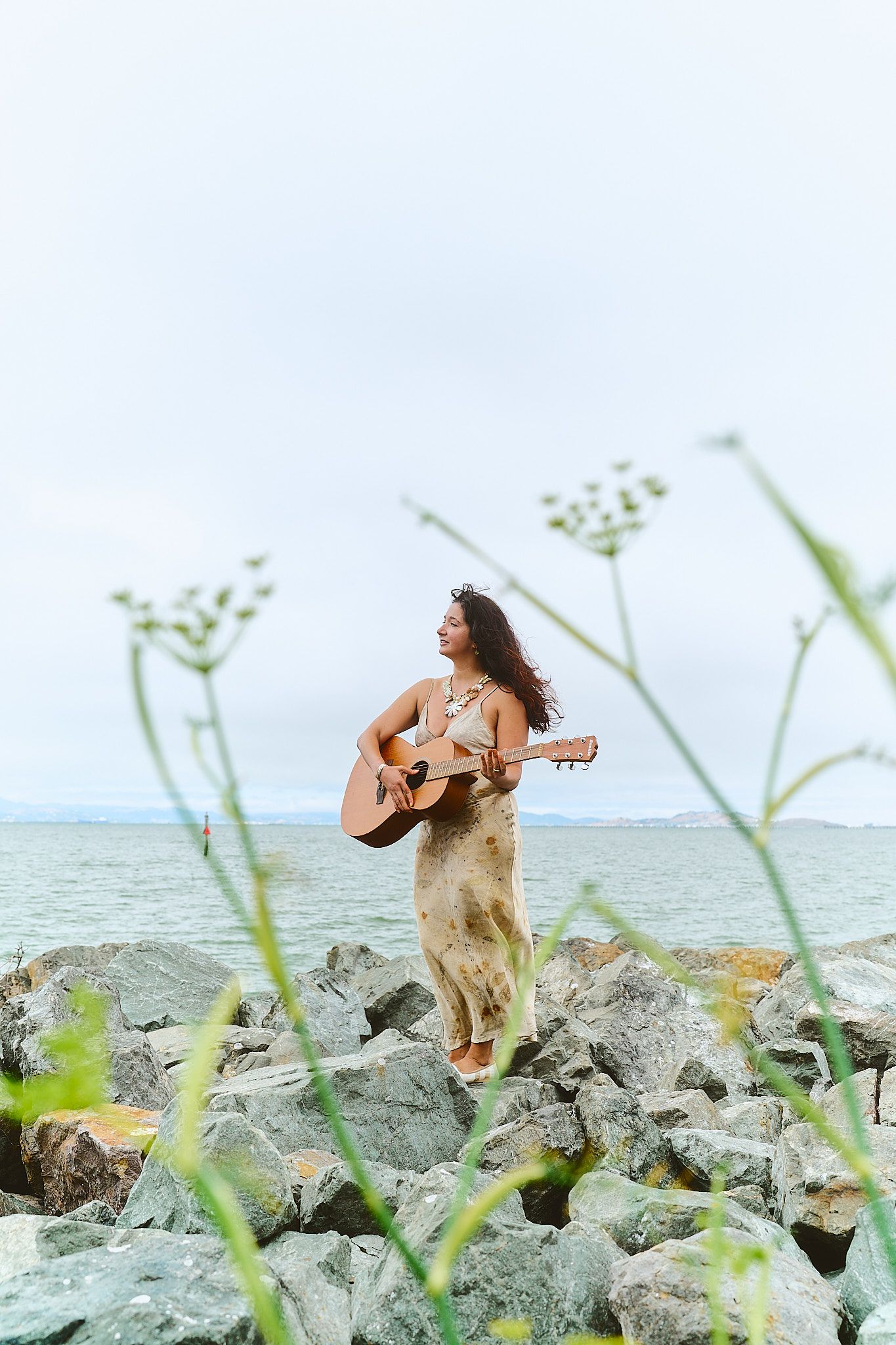 Birthday editorial photo shoot of a woman in flowy dress playing guitar while standing on rocky Bay Area coastal shoreline.