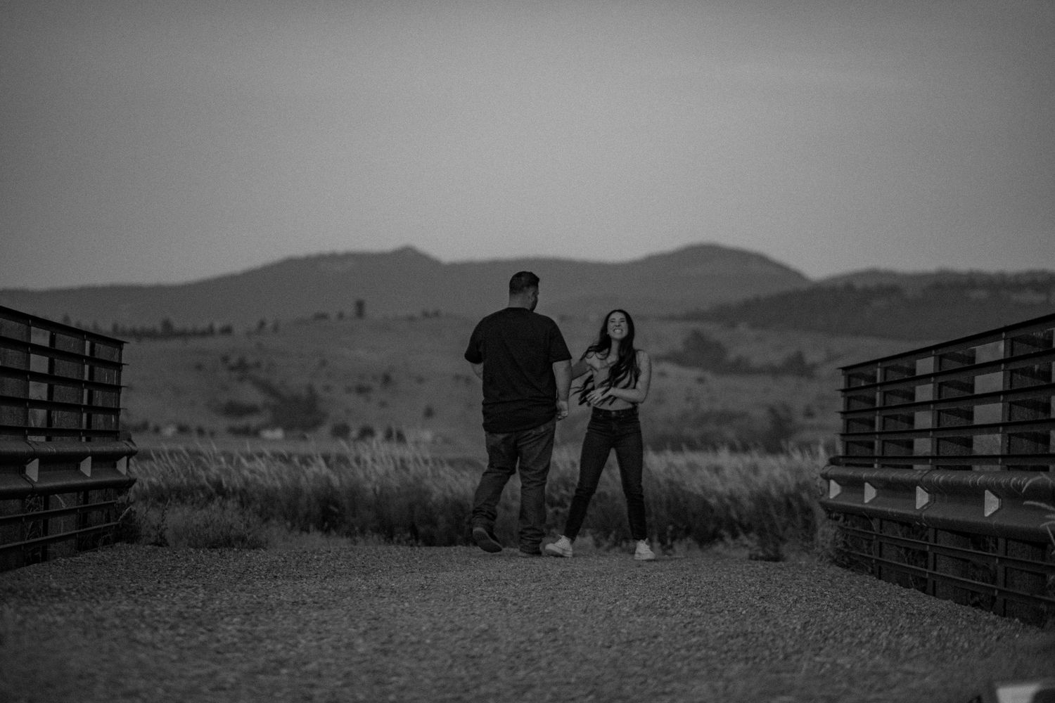 A couple stands together on a wooden boardwalk overlooking rolling hills and mountains in black and white.