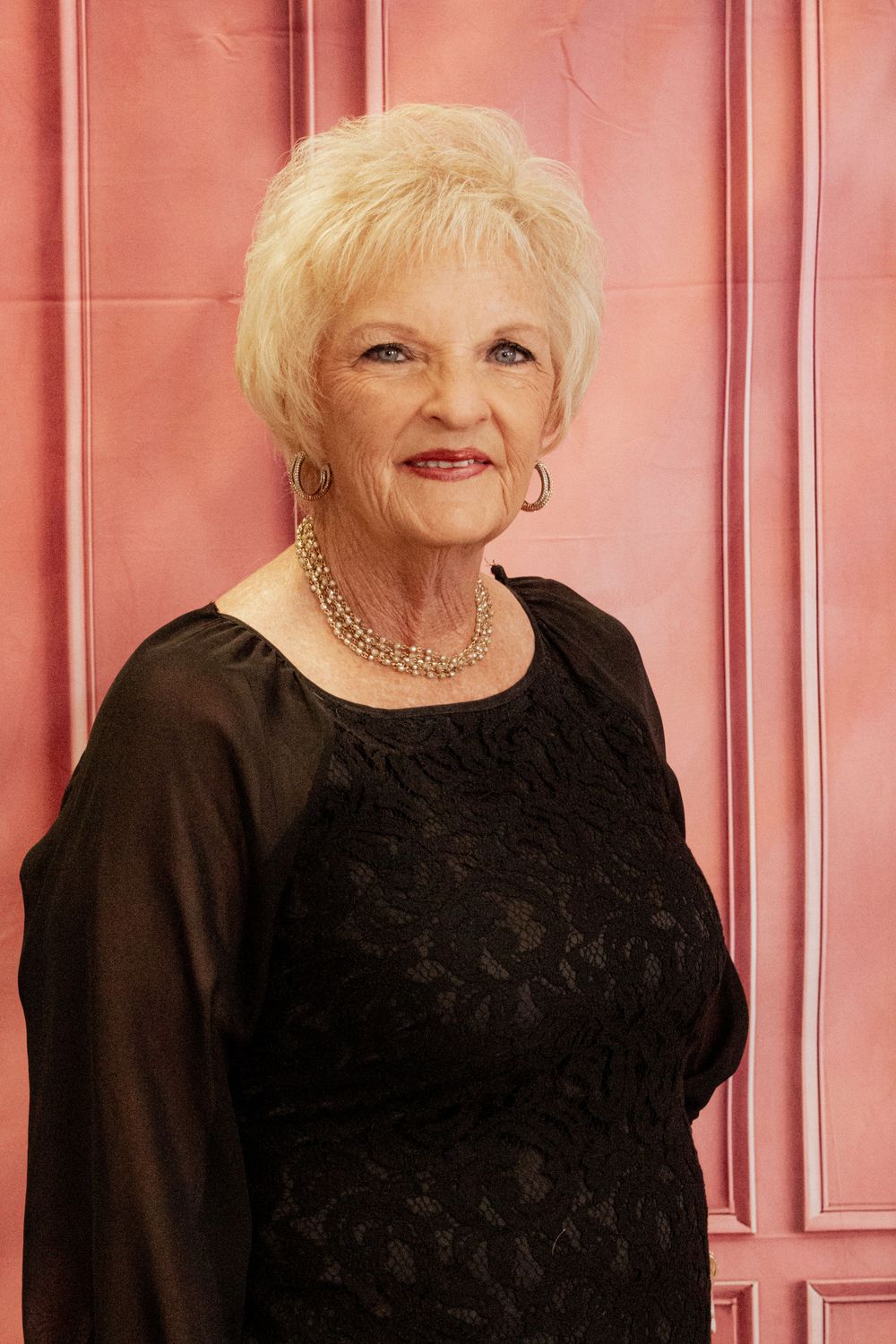 A person with platinum blonde hair wearing a black top poses in front of a pink paneled wall.