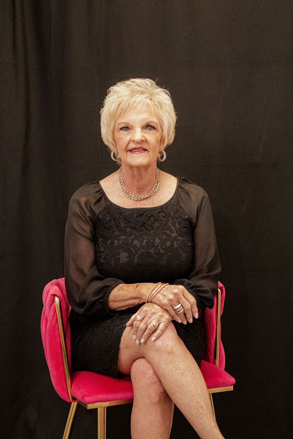 A woman in a black dress and sheer sleeves sits elegantly on a bright pink chair against a dark backdrop.