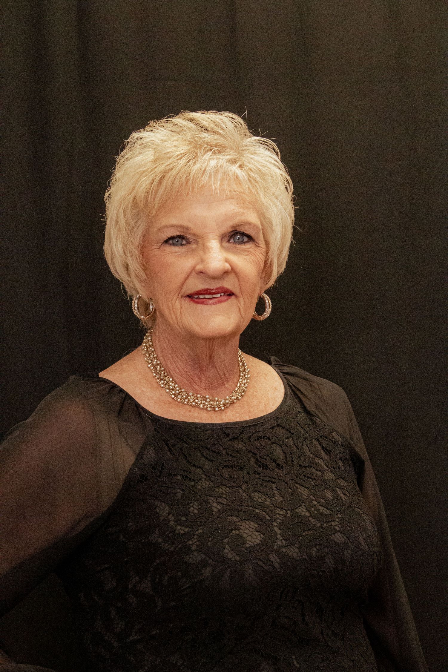 Portrait shots of a person with short blonde hair wearing a black lace top and smiling warmly against a dark background.