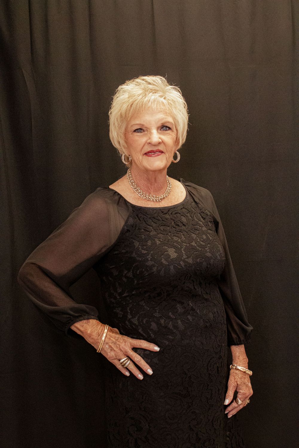 Senior woman wearing elegant black dress with sheer sleeves poses against dark background in series of glamour photos.