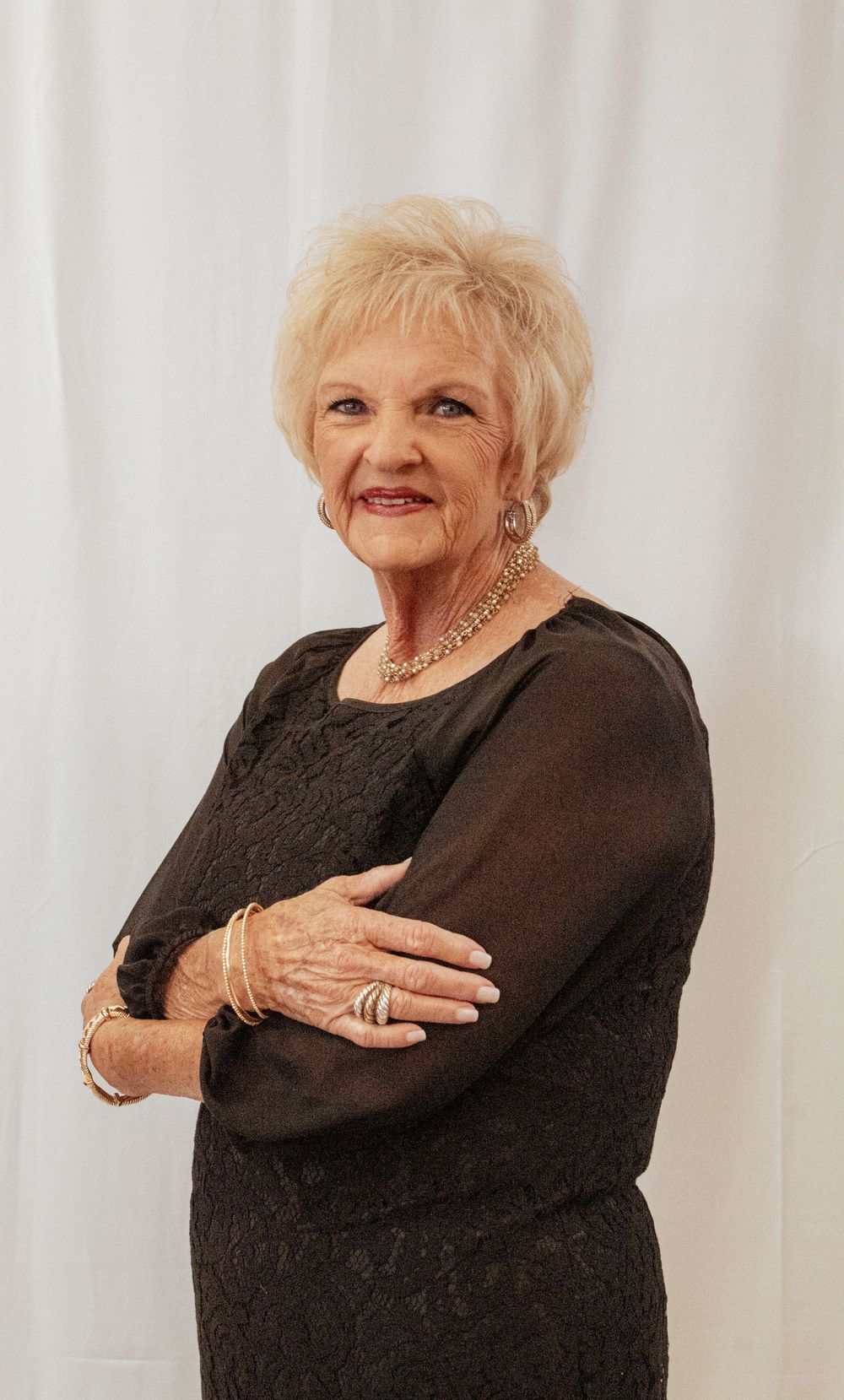 Senior woman in black lace dress poses against white backdrop wearing jewelry and a warm smile.