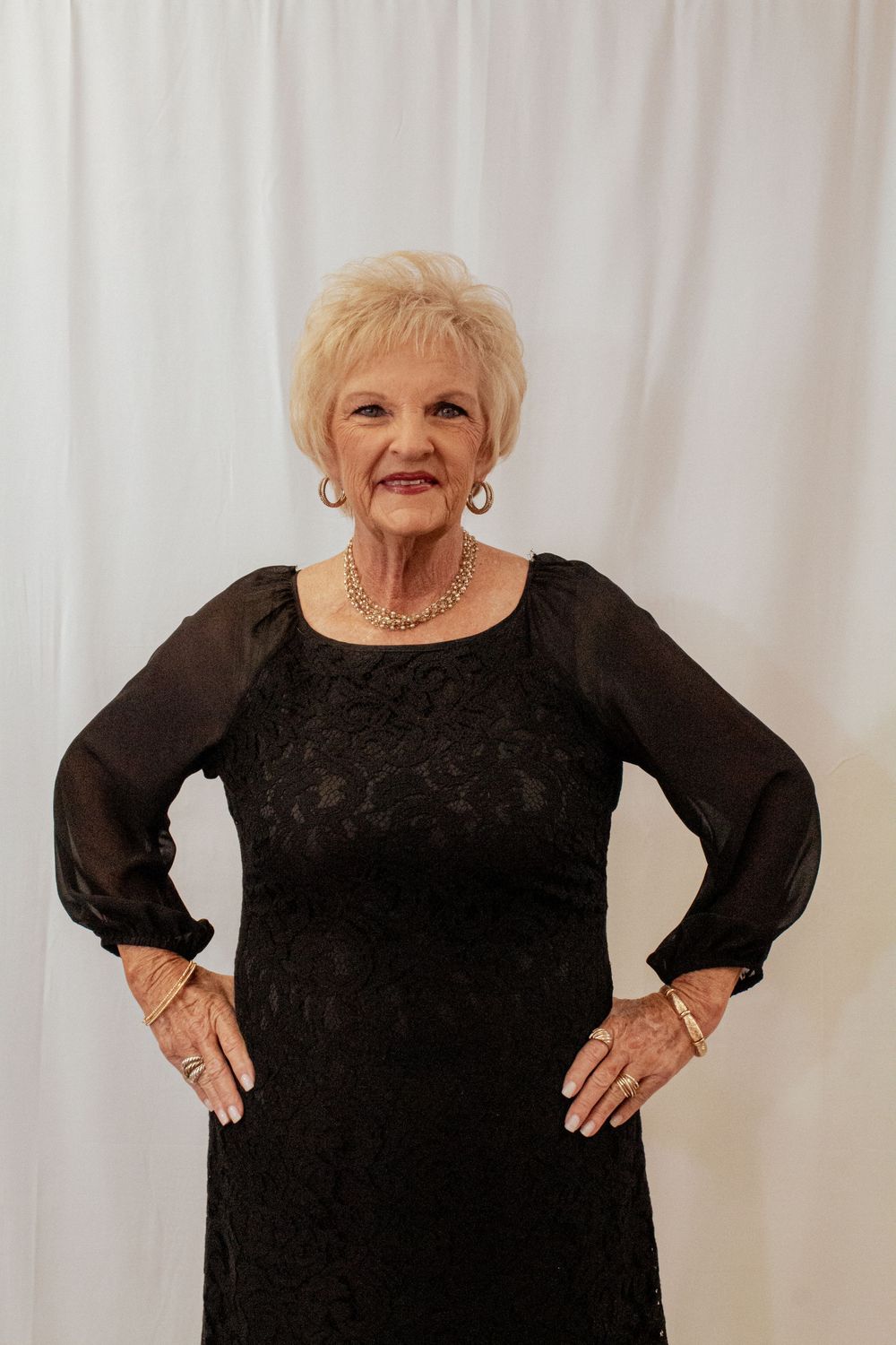 An elegant older woman poses in a long-sleeved black velvet dress against a white backdrop.