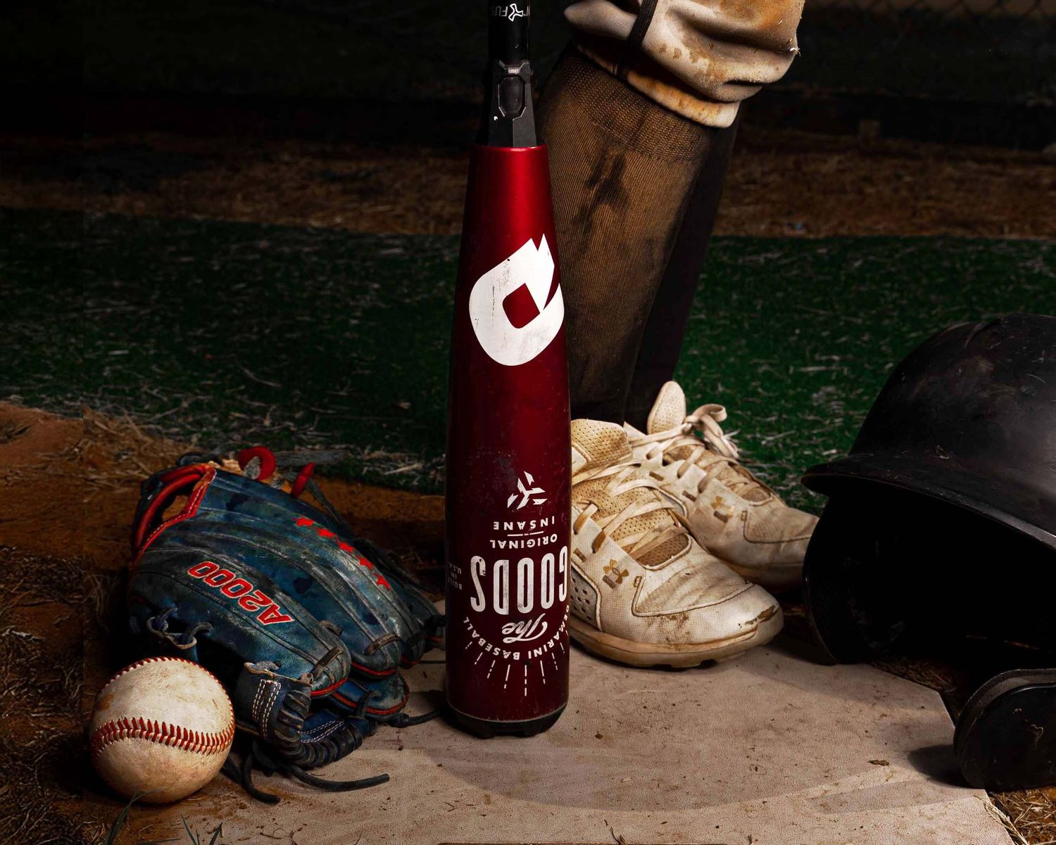A baseball still life with red bat, glove, ball and cleats arranged on artificial turf against a dark background.