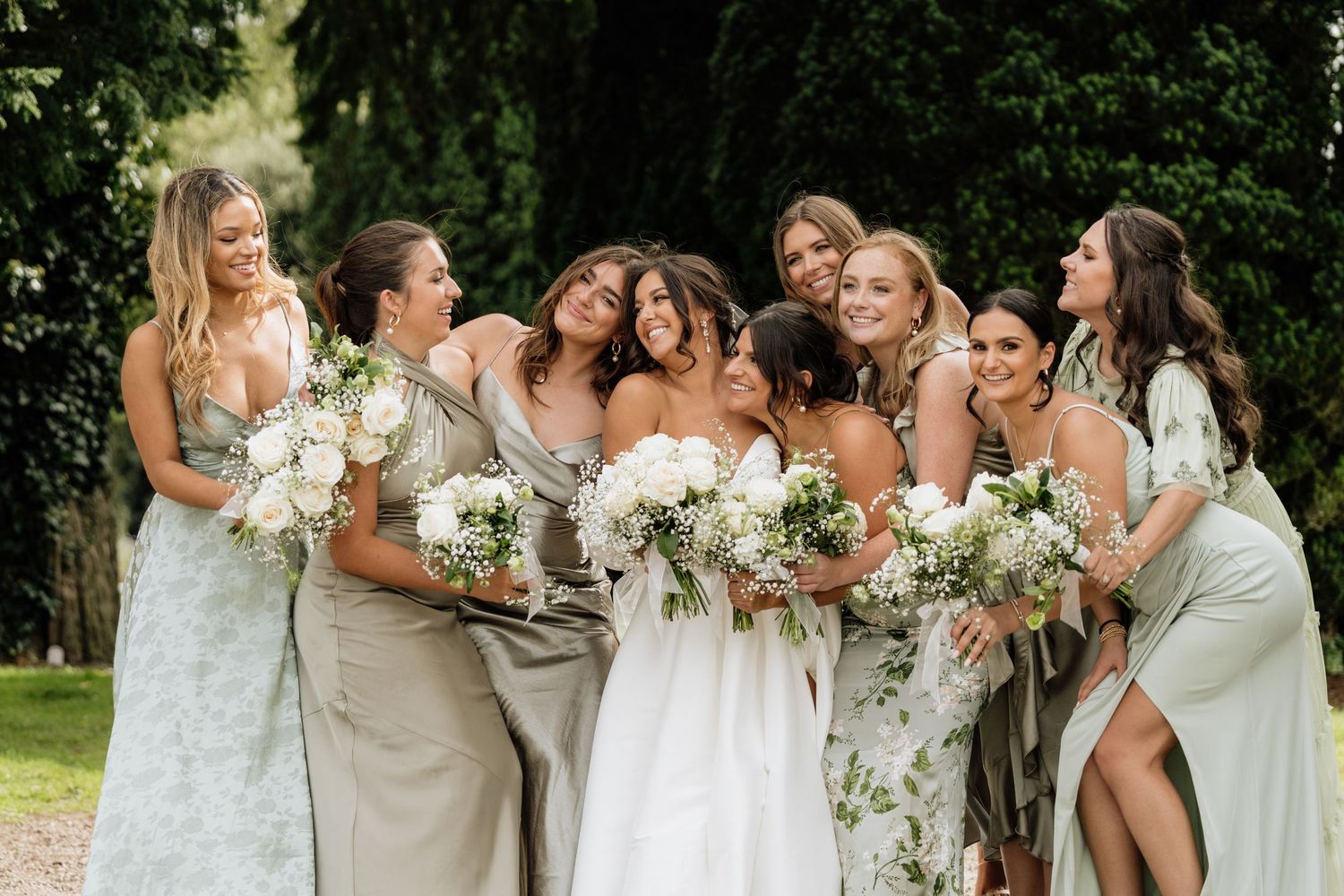 A bridal party in light-colored dresses poses together with white bouquets.