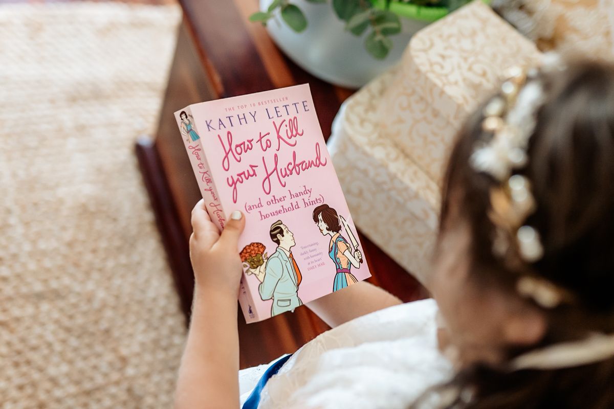 A hand holds a pink romance novel against a beige fabric background.