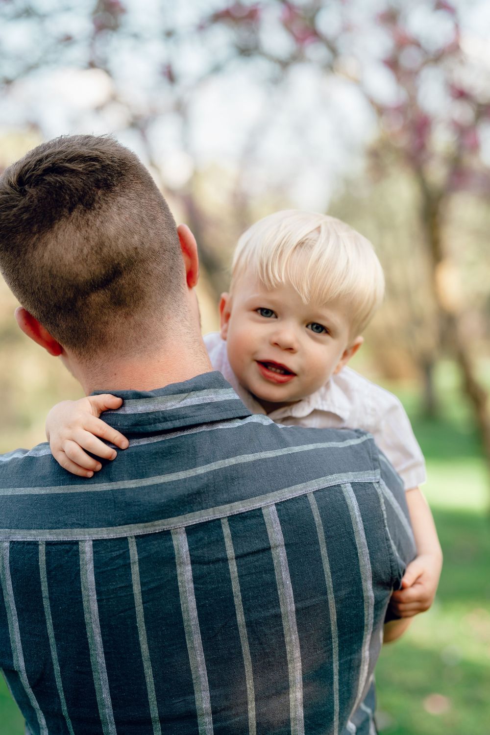 Toddler with blonde hair peeks over daddy's shoulder while being carried outdoors in a spring garden at F.A. Seiberling Nature Realm, Akron