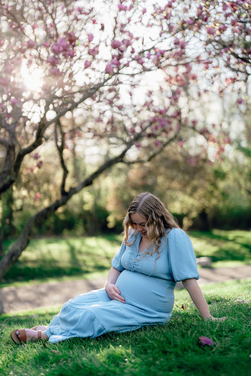 A pregnant woman in a blue dress sits peacefully on grass beneath blooming spring trees at maternity session at F.A. Seiberling Nature Realm