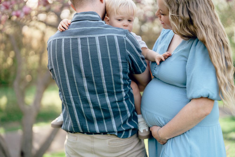 A pregnant woman in a blue dress stands with a family member holding a small child in a F.A. Seiberling Nature Realm Maternity session.