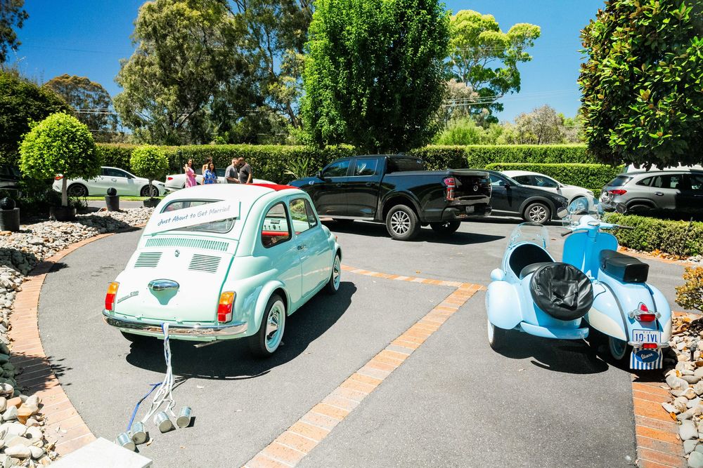 Vintage blue classic car parked in sunny parking lot surrounded by green trees and other vehicles.
