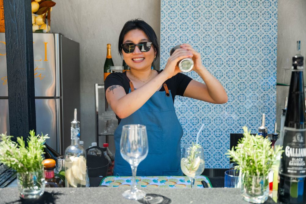 A bartender in a blue top prepares cocktails behind a counter decorated with fresh herbs and glassware.