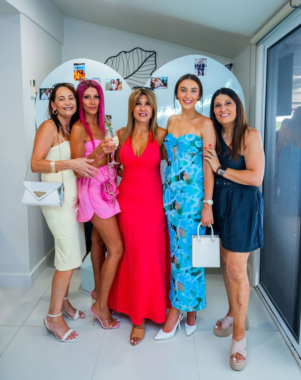 A group of friends pose together in a stylish beachfront event venue wearing colorful summer dresses and sandals.