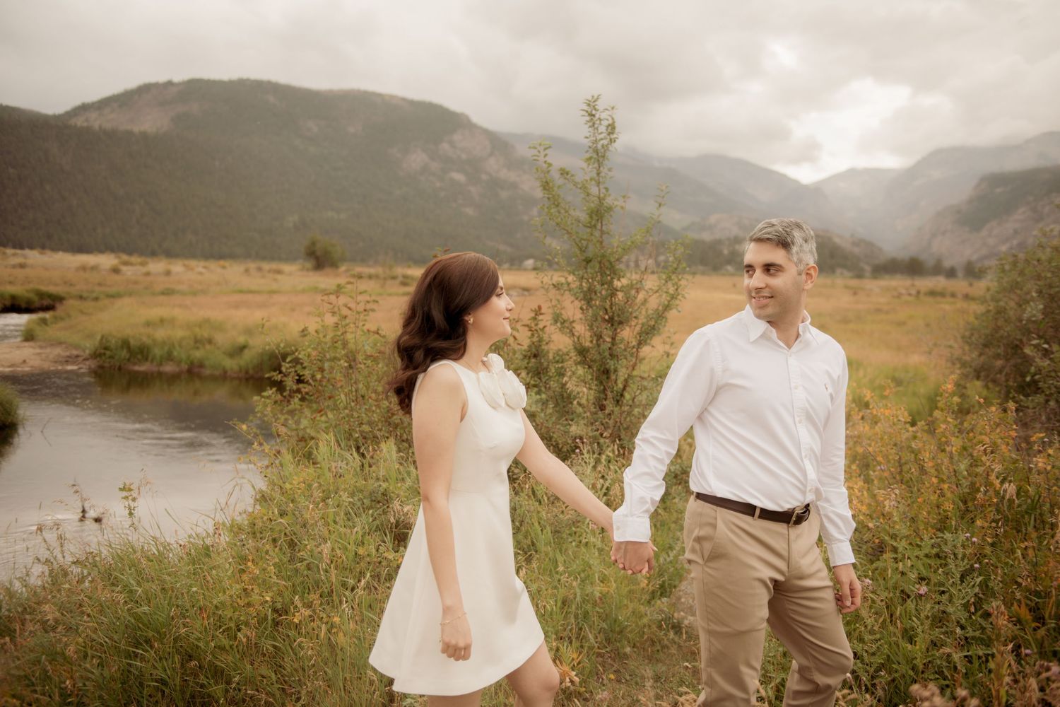 Two people hold hands while standing in a grassy mountain meadow beside a river on an overcast day.