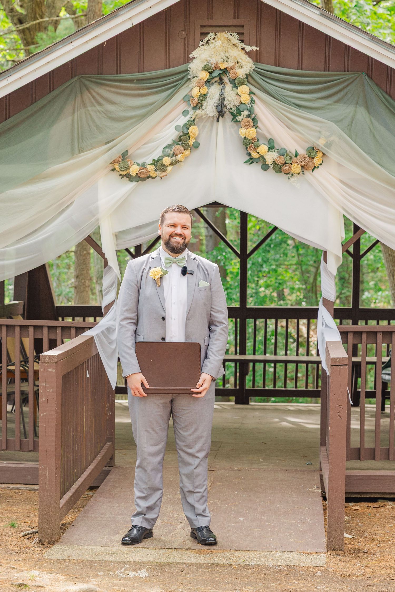 Wedding ceremony gazebo decorated with white drapes and floral garlands while someone stands beneath holding a folder.