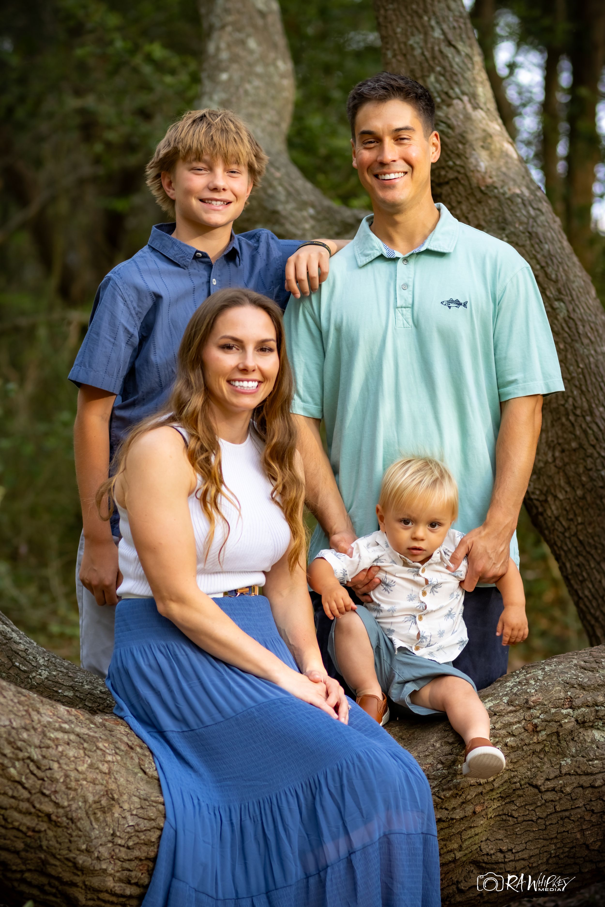 A family poses together on a tree branch outdoors, wearing coordinated blue and white casual outfits.
