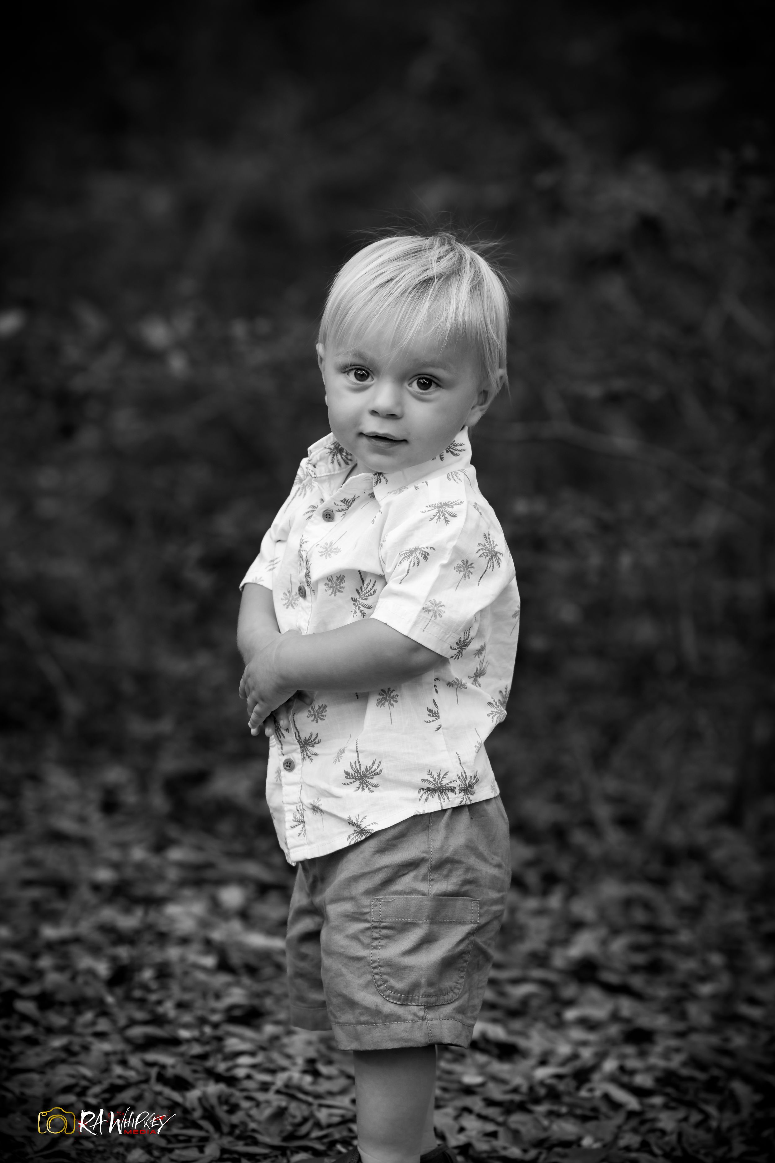 Black and white portrait of a small child in floral shirt and shorts standing outdoors on fallen leaves.