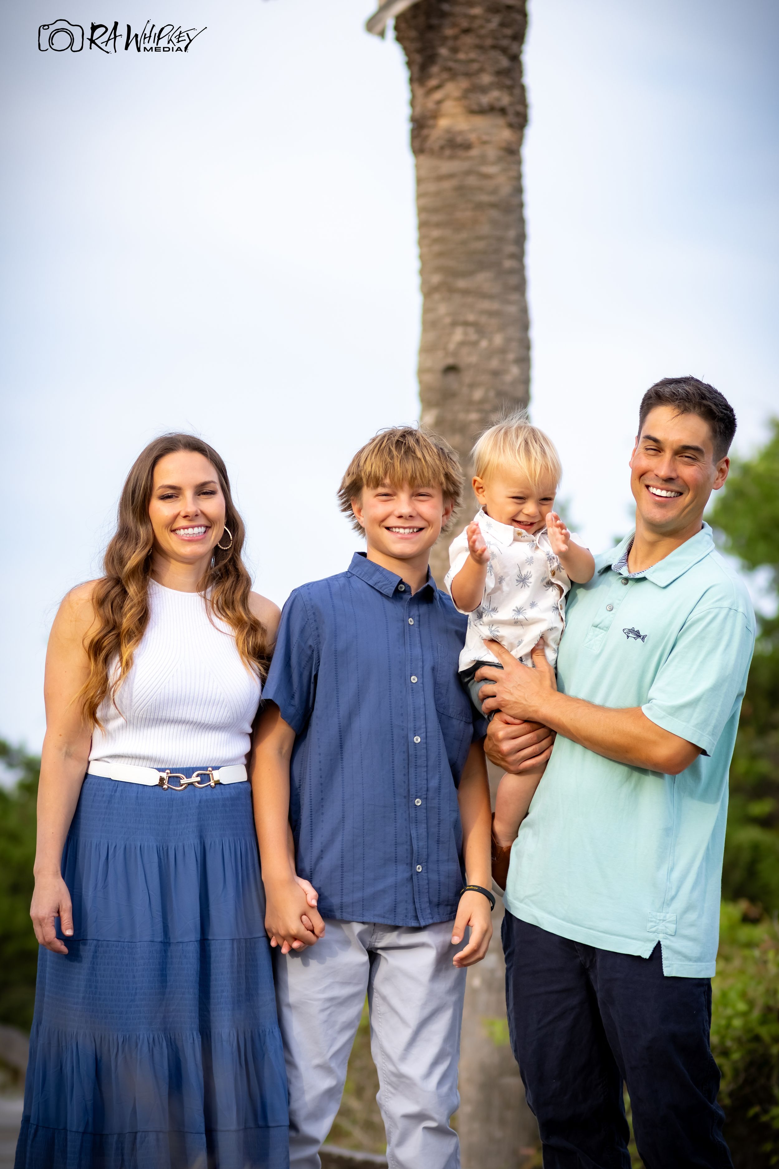 A group stands together outdoors wearing coordinating blue and white outfits with a palm tree trunk visible in the background.