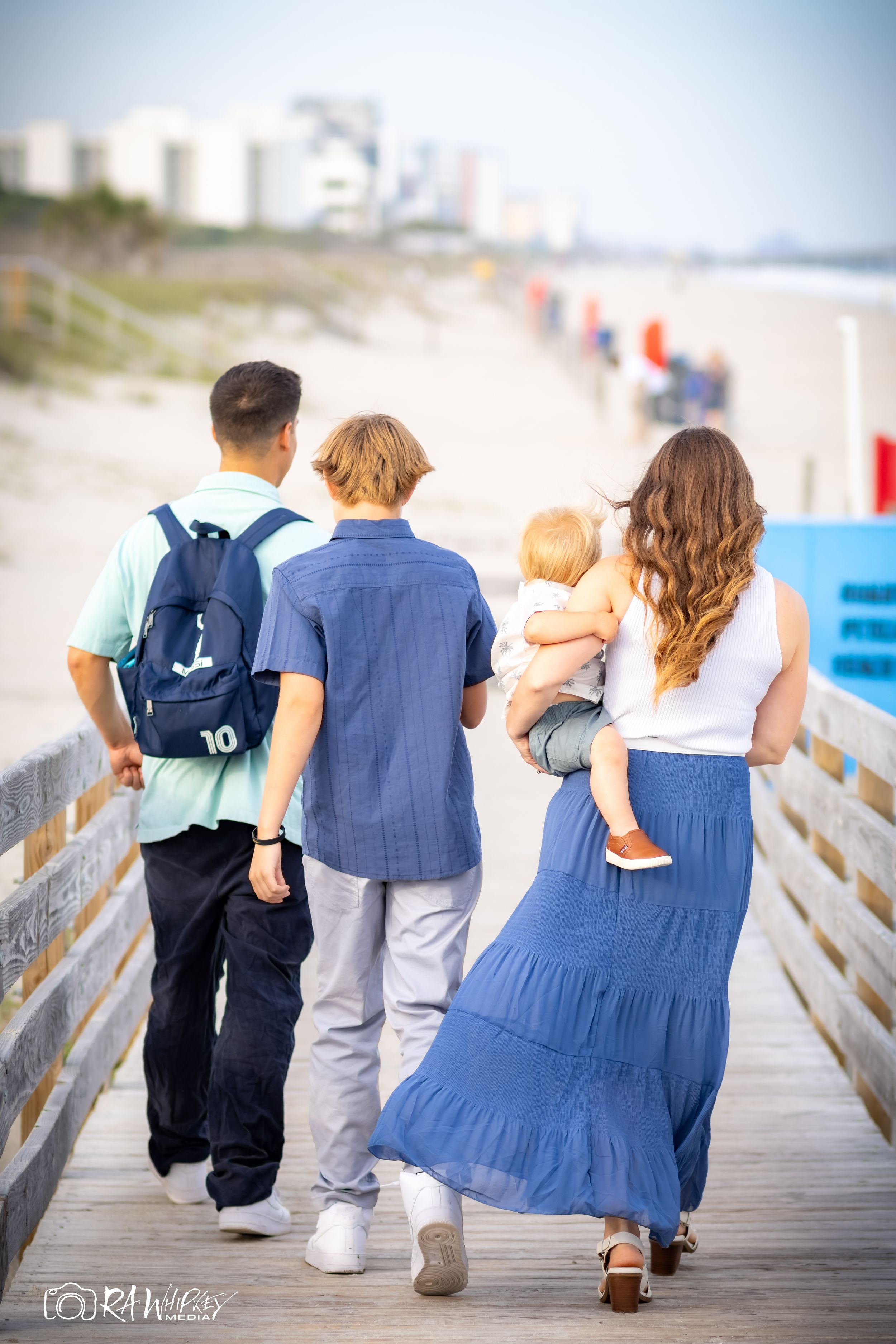 A family walks together on a wooden boardwalk along a beach with coastal buildings visible in the background.