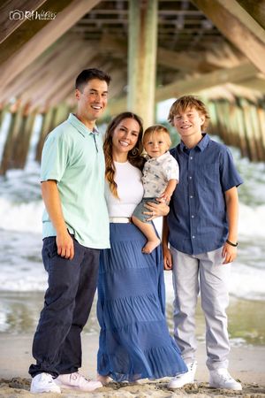 Family of four poses together under a pier at the beach wearing coordinated blue and white casual outfits.