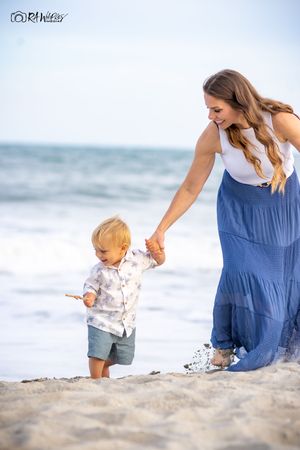 A tender moment captured at the beach as parent in blue dress holds little one's hand while walking on sandy shore by ocean.