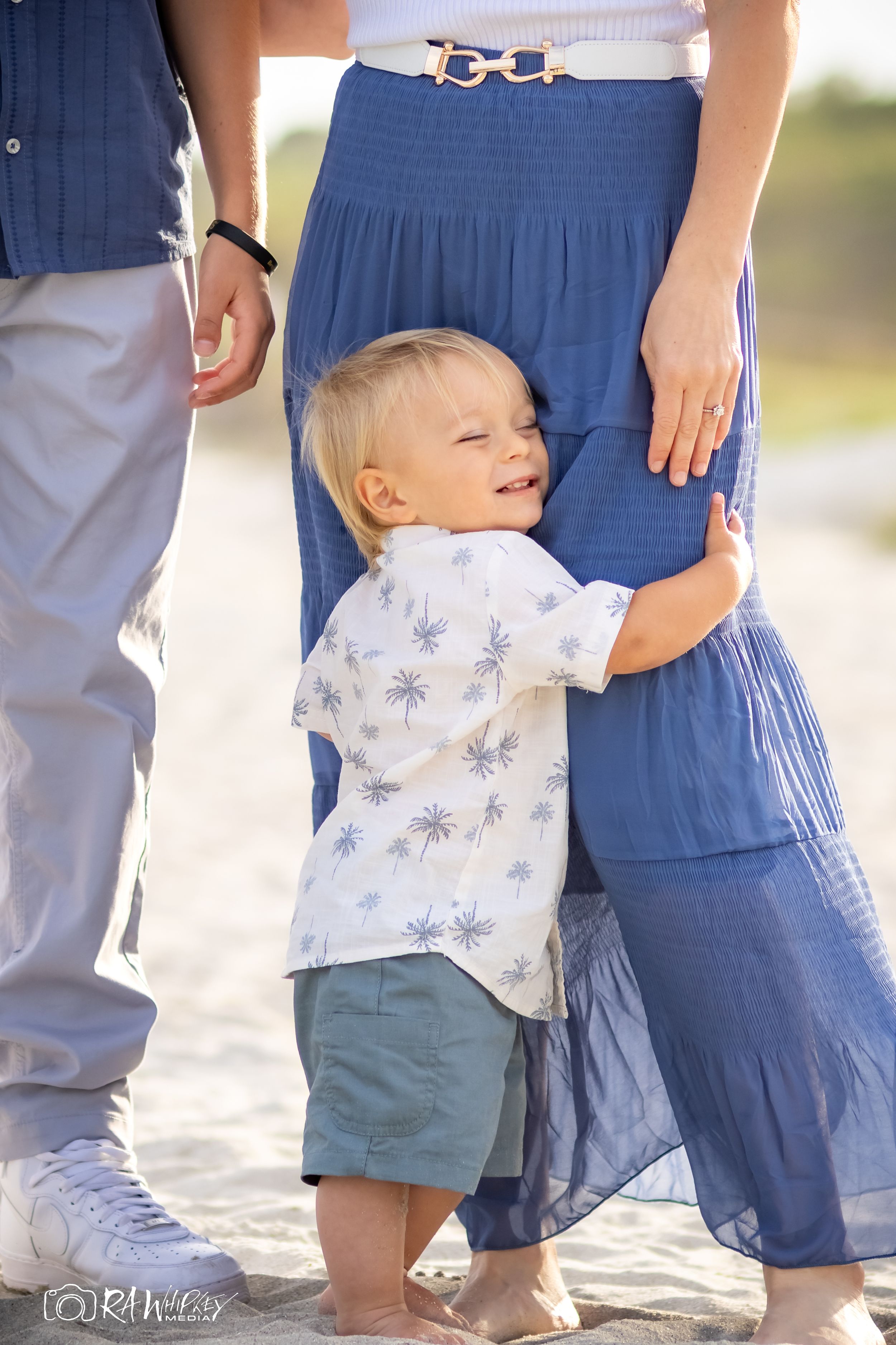Young child in palm print shirt and shorts hugs a person wearing a blue maxi skirt on a sunny day.