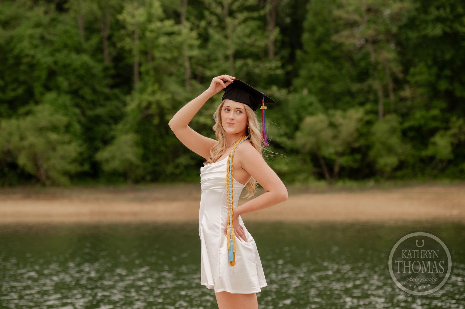 A person in a white sundress and sun hat poses confidently by a serene lake with trees in the background.